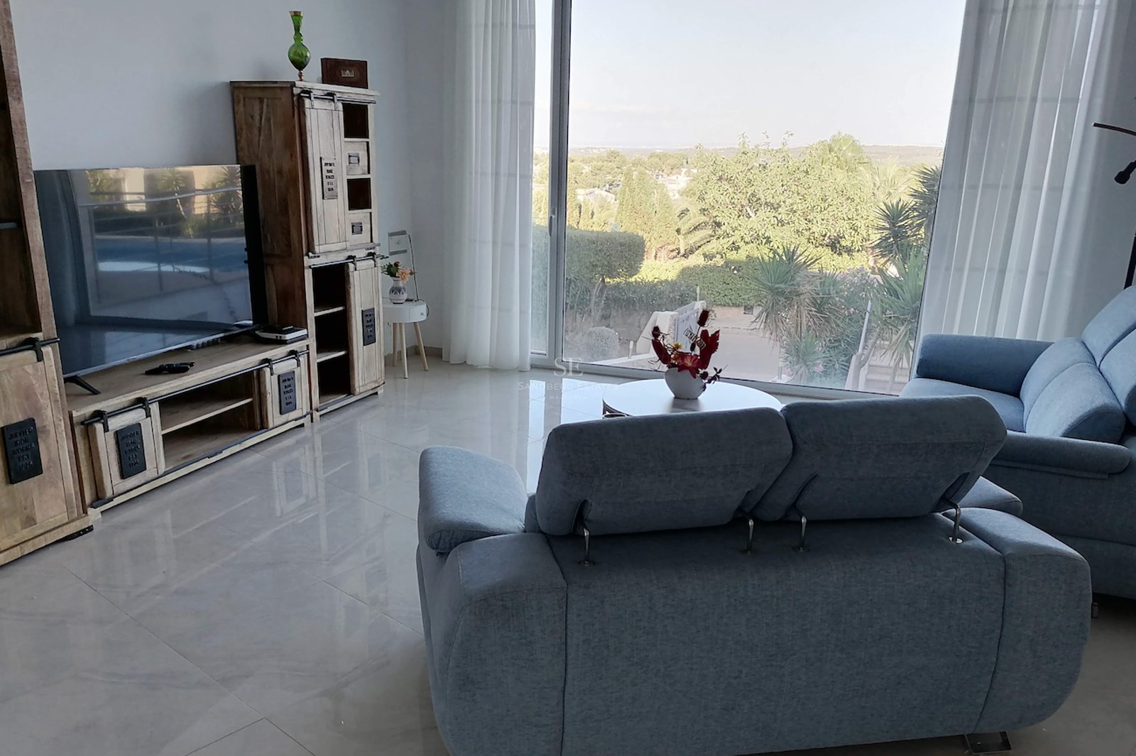 Modern living room featuring grey fabric sofas, polished floor tiles, and a large window overlooking lush greenery.