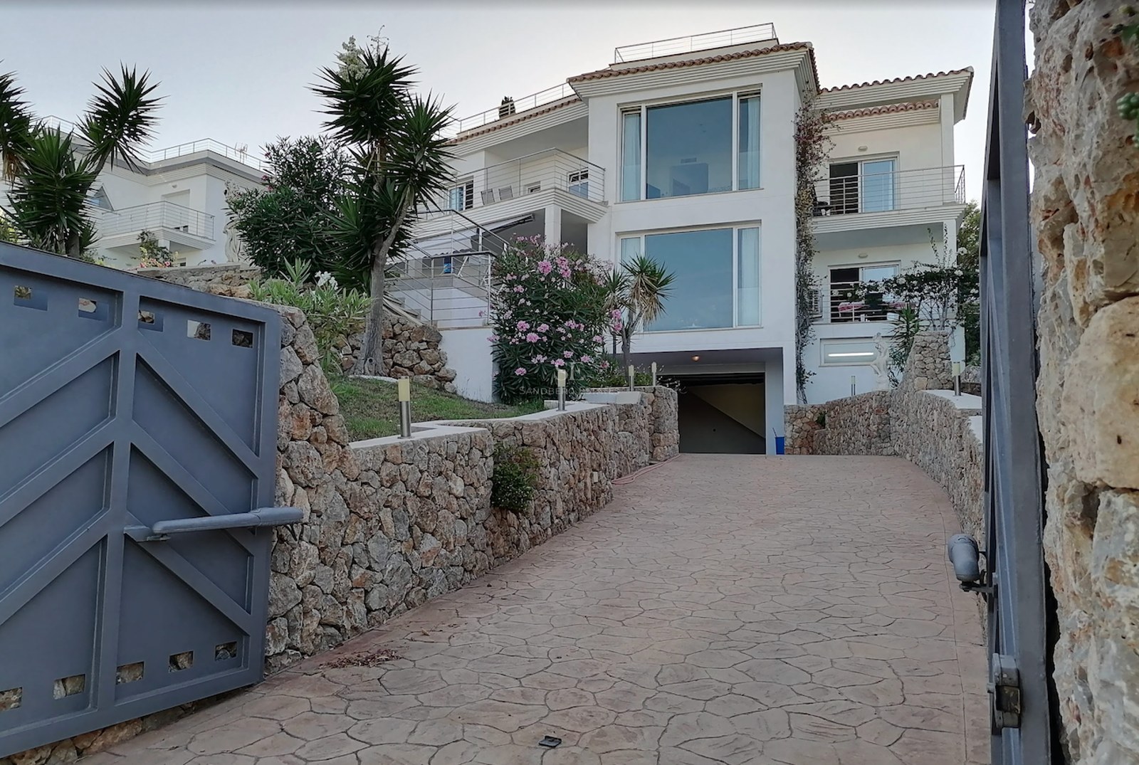 View of a multi-story white villa with large windows, seen from a gated driveway lined with natural stone walls.