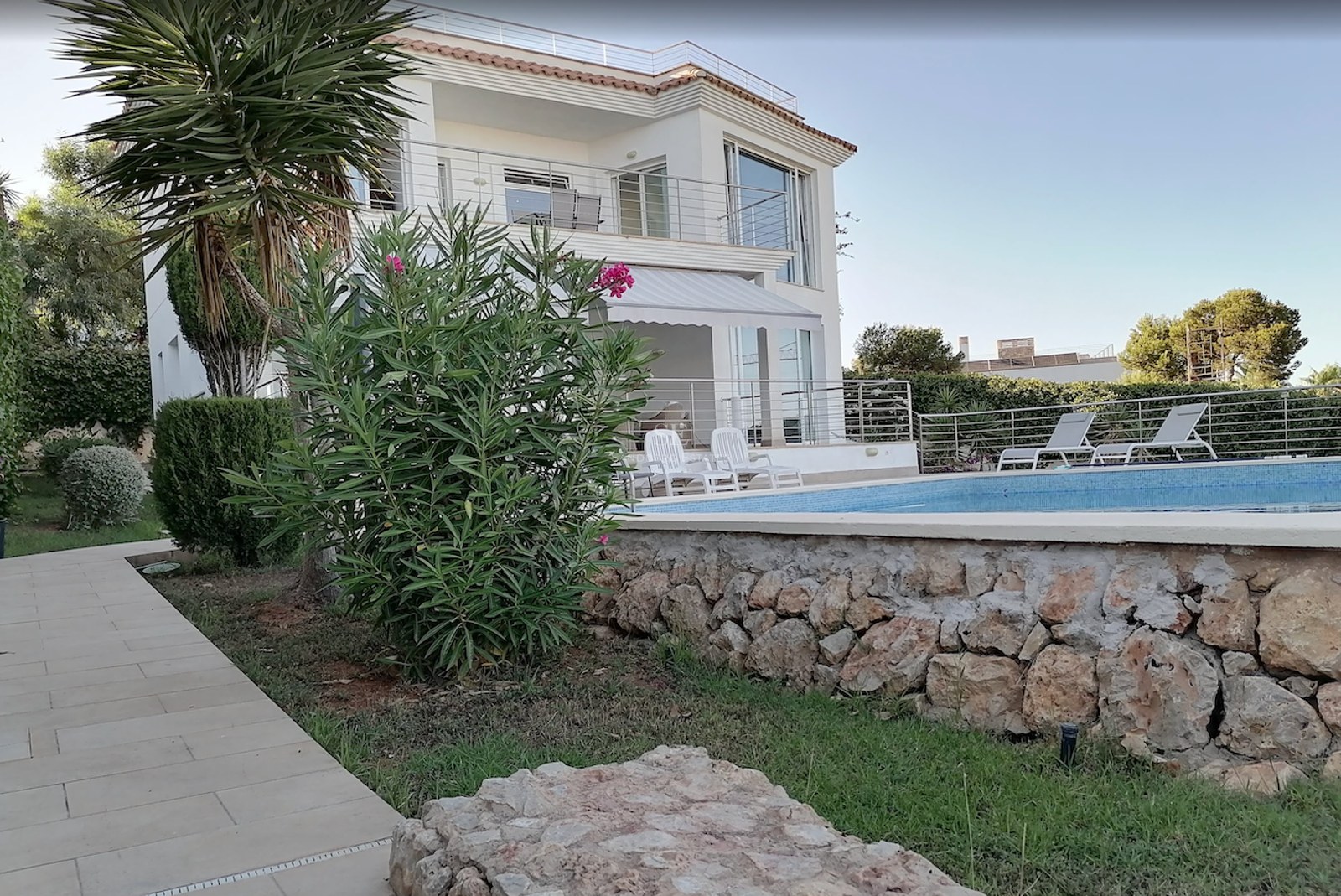 White two-story villa overlooking a turquoise pool with sun loungers and a natural stone garden wall.
