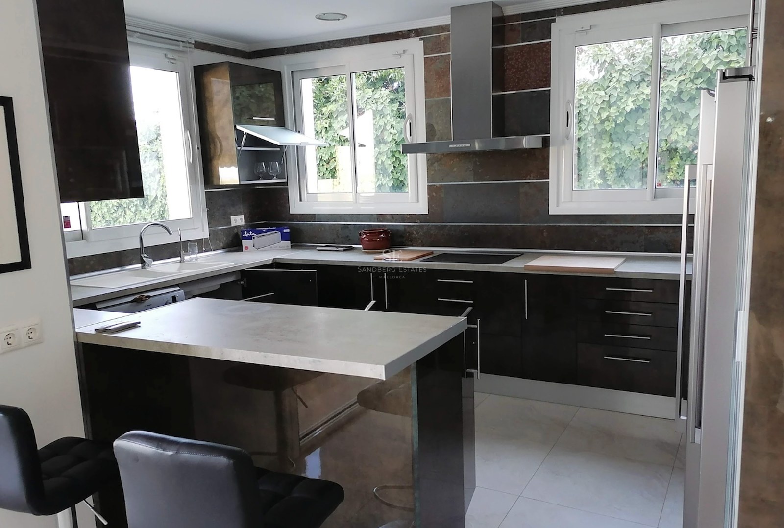 Modern L-shaped kitchen with dark glossy cabinetry, grey countertops, and windows overlooking lush greenery.