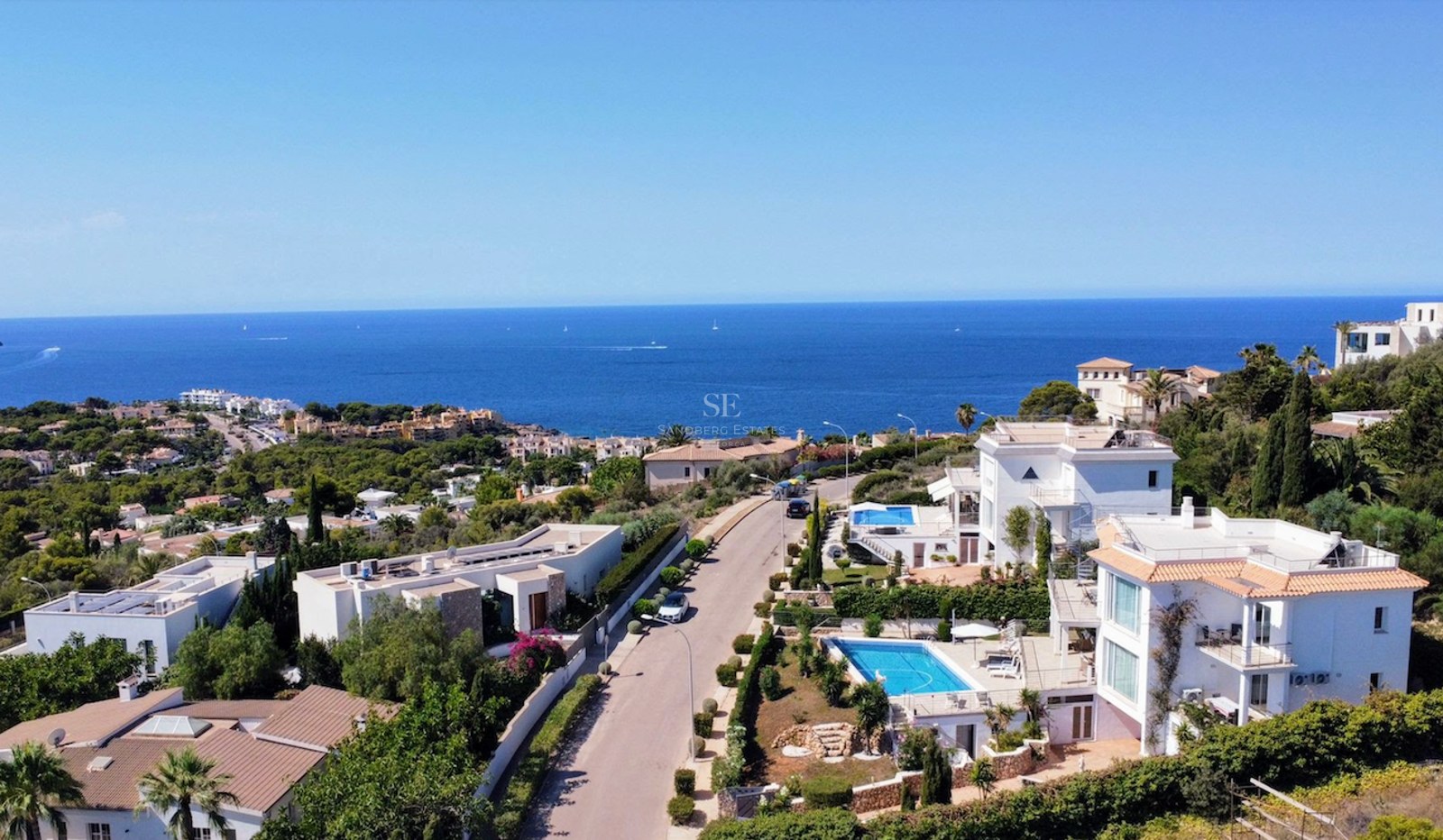 Aerial view of luxury white villas with swimming pools overlooking the deep blue Mediterranean Sea under a clear sky.