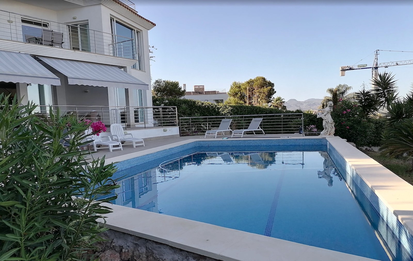 A rectangular blue mosaic-tiled swimming pool in front of a white luxury villa with sun loungers and awnings.
