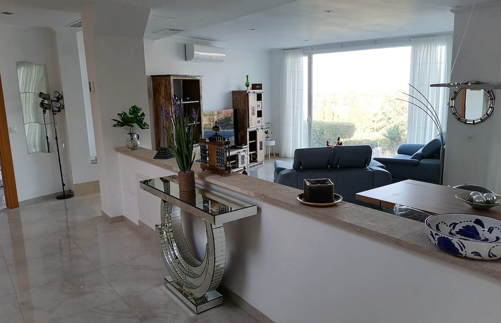 Modern living room featuring marble floors, blue seating, and a large window overlooking lush greenery.