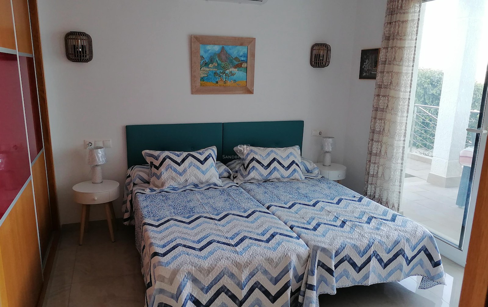 Bedroom with twin beds, teal headboard, white walls, and floor-to-ceiling glass door leading to a sunny terrace.