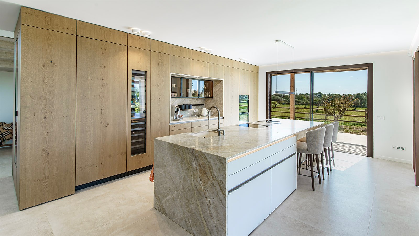 Modern kitchen featuring a marble waterfall island, oak cabinetry, and floor-to-ceiling windows overlooking a garden.