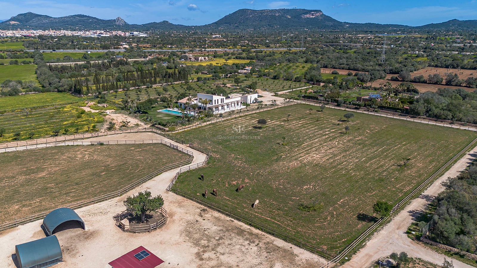 Aerial view of a large white modern villa with a swimming pool and vast green paddocks against a mountain backdrop.