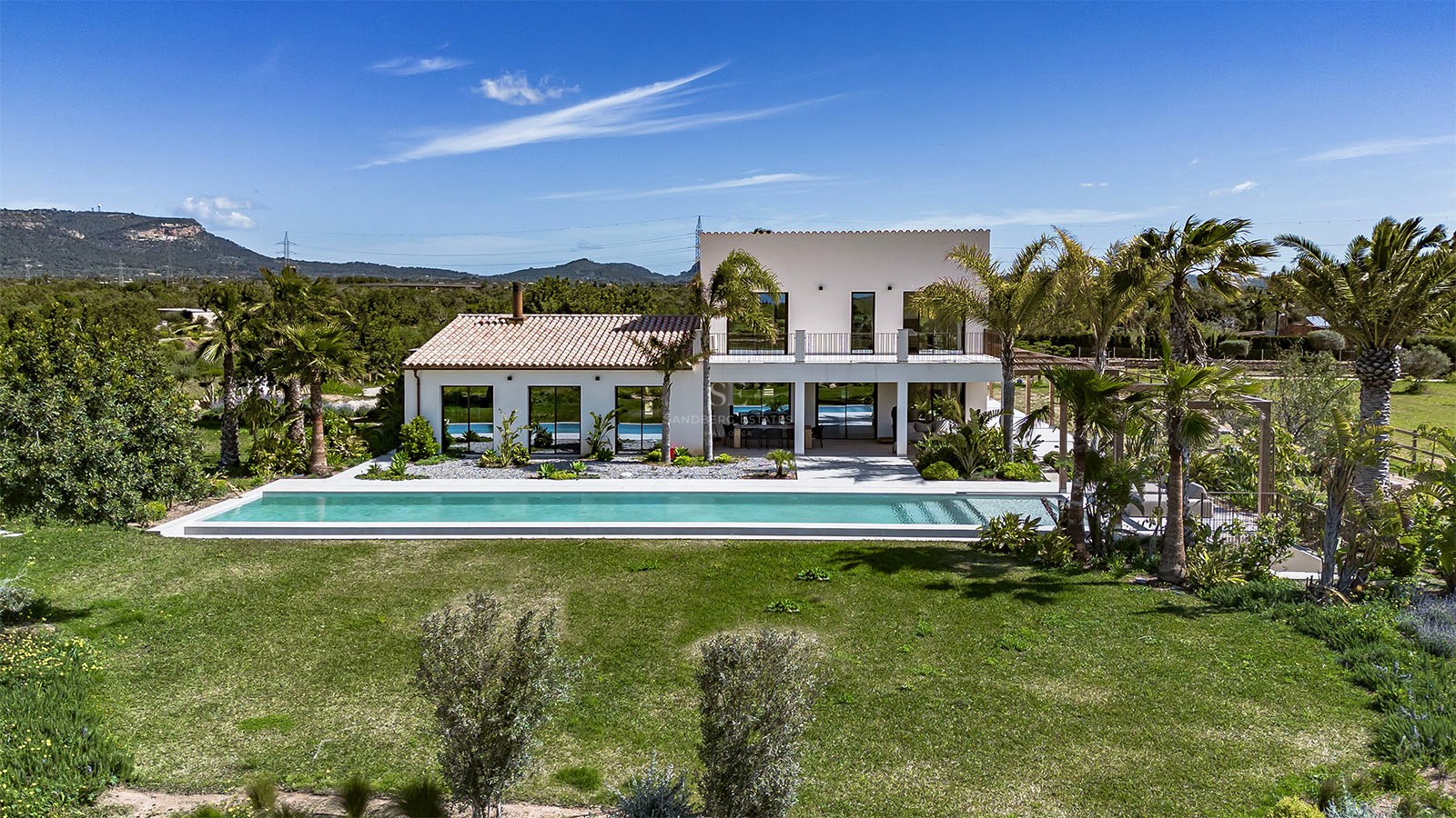 Rear view of a white two-story villa with a long swimming pool, green lawn, and mountain backdrop under a blue sky.