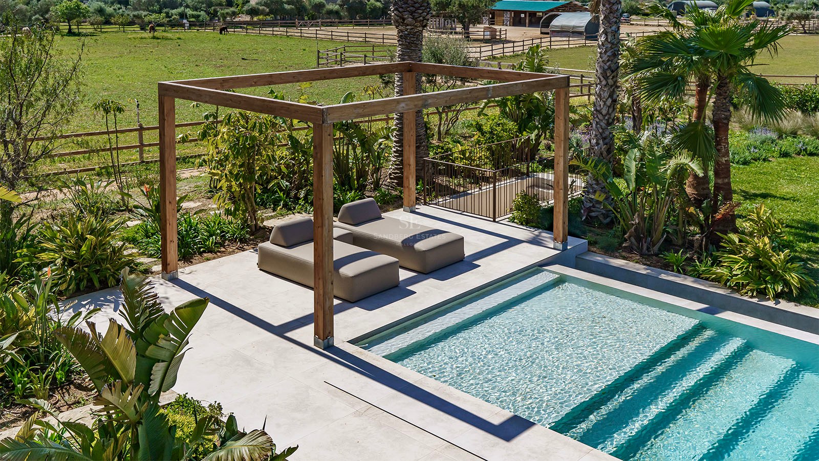 Two modern sunbeds under a wooden pergola beside a turquoise pool overlooking a green field.