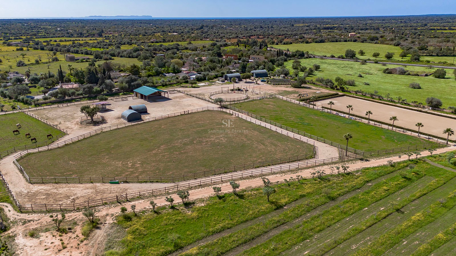 Aerial shot of fenced horse paddocks, stables, and green fields with the Mediterranean sea on the horizon.