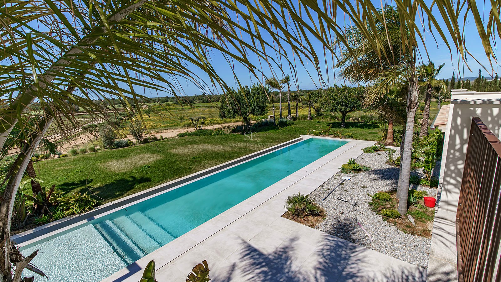 A long turquoise lap pool surrounded by white stone decking and a lush green lawn under a clear blue sky.