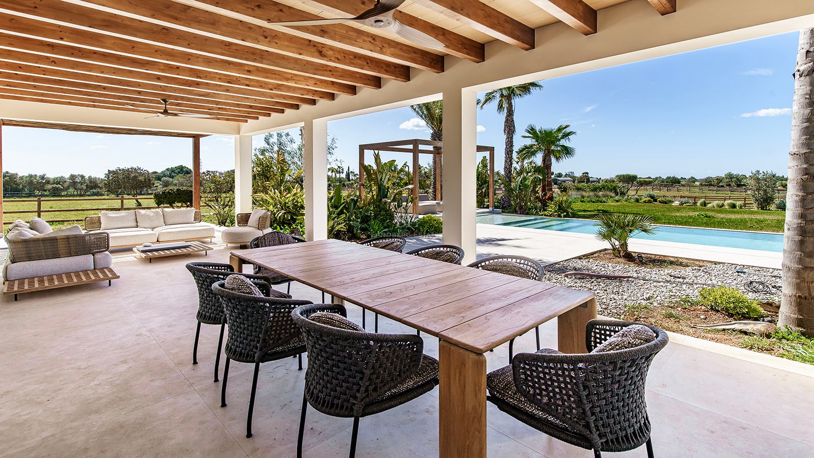 Wooden dining table and woven chairs on a covered terrace with exposed timber beams overlooking a pool and garden.