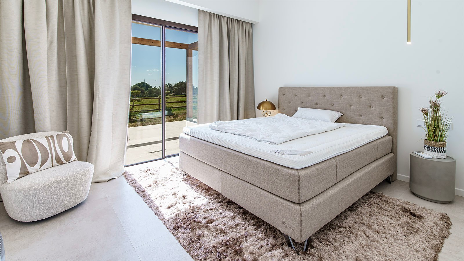 High-end bedroom featuring a tufted bed, plush rug, and floor-to-ceiling glass doors opening onto a sunny terrace.