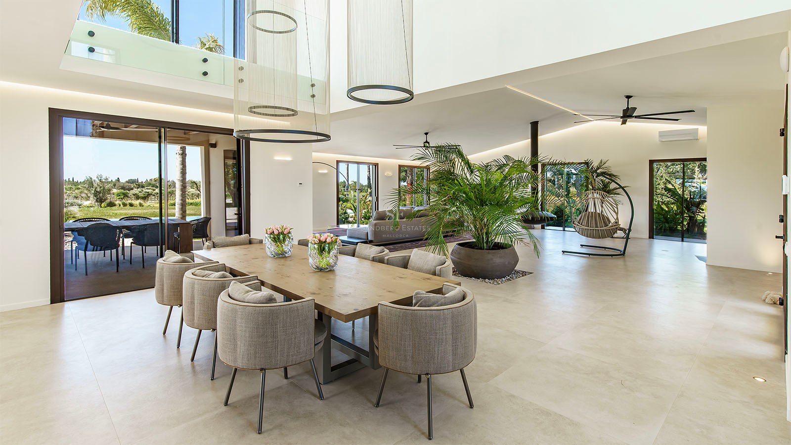 Modern open-plan dining room with a large wooden table, grey chairs, high ceilings, and floor-to-ceiling glass doors.