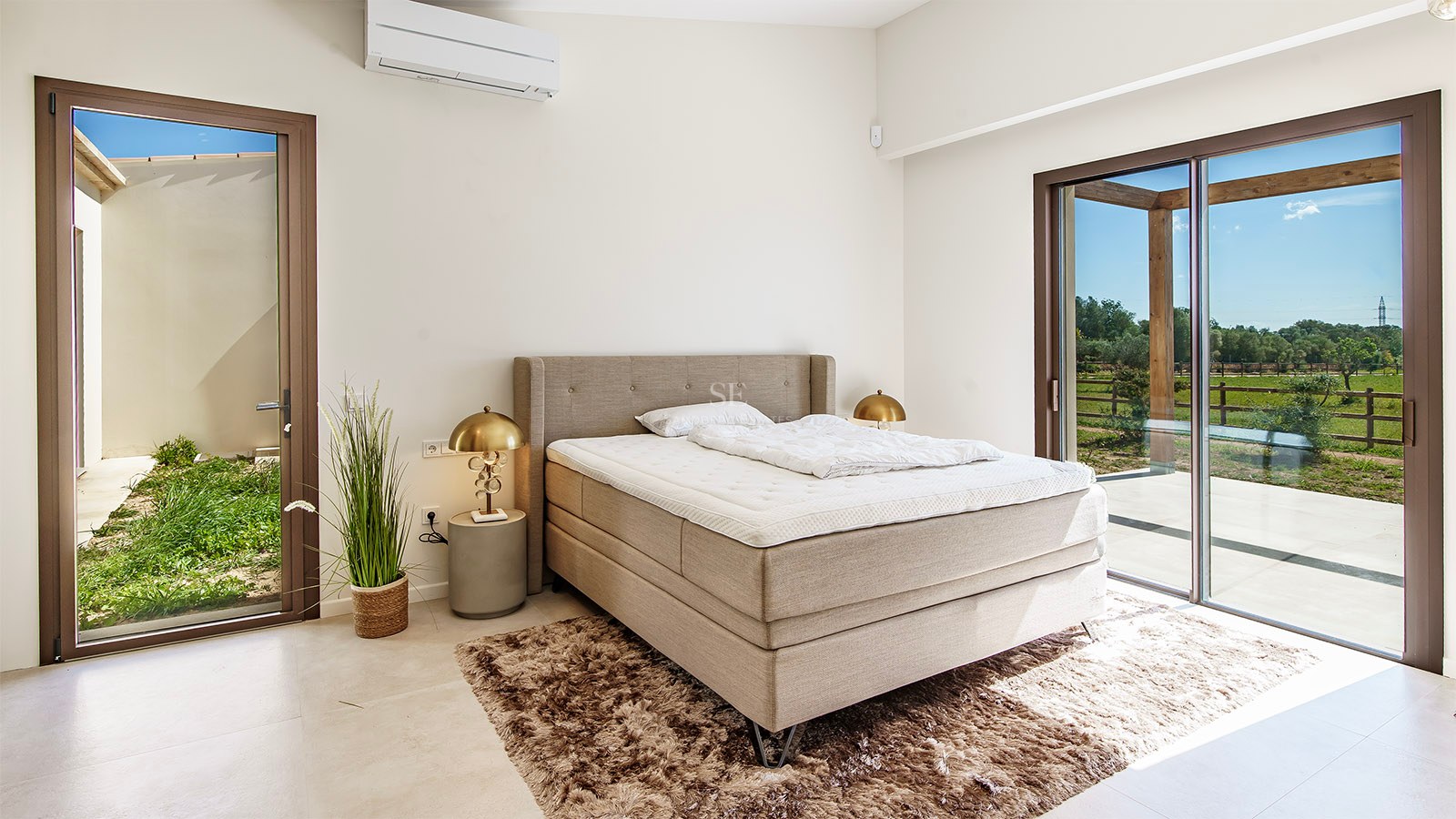 Bright bedroom featuring a large upholstered bed, gold bedside lamps, and a glass door opening to a garden patio.