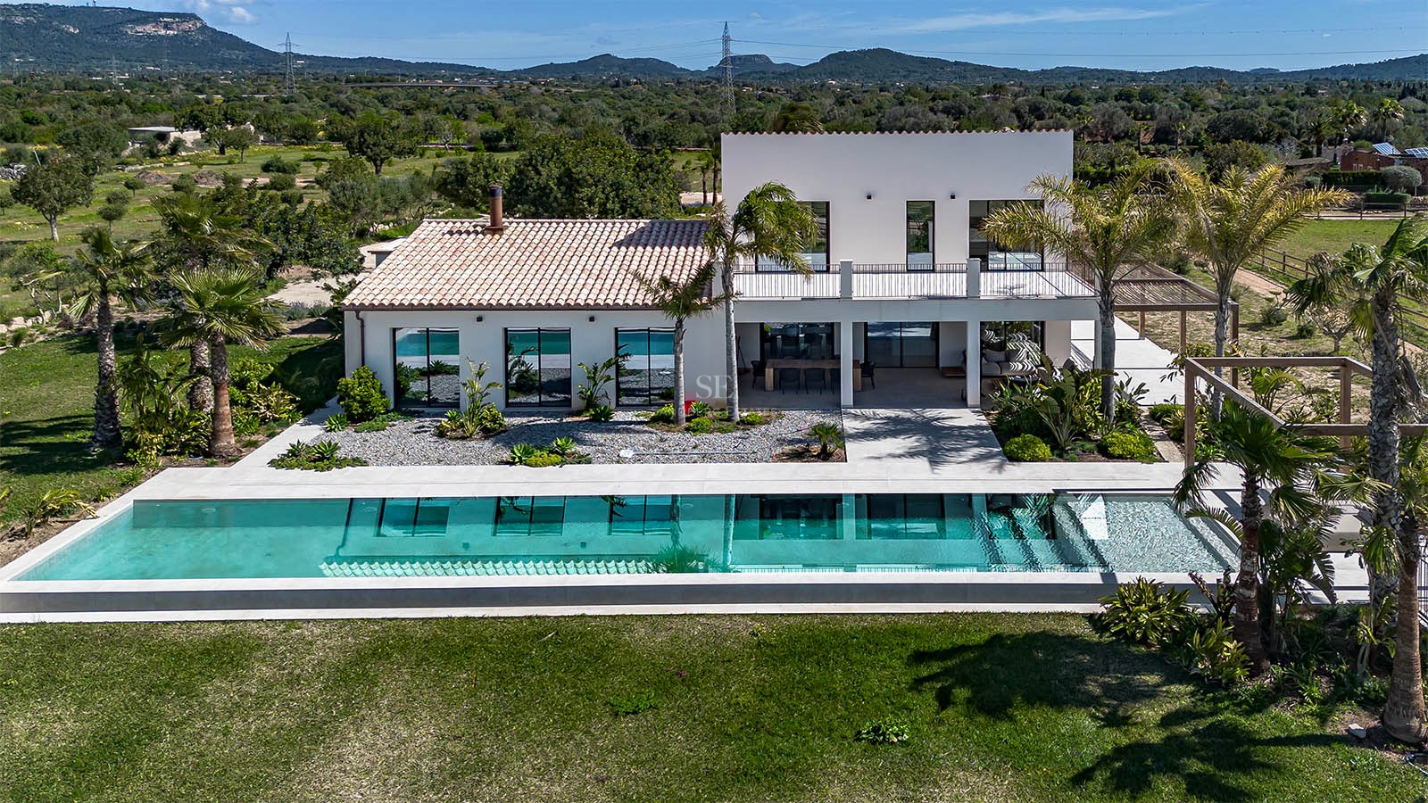 Aerial view of a modern white villa with a turquoise swimming pool and mountains in the background.