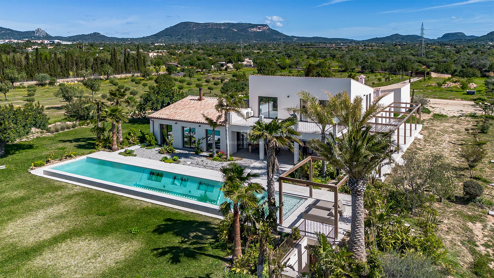 Aerial view of a white luxury villa featuring a long rectangular pool, palm trees, and mountains in the distance.