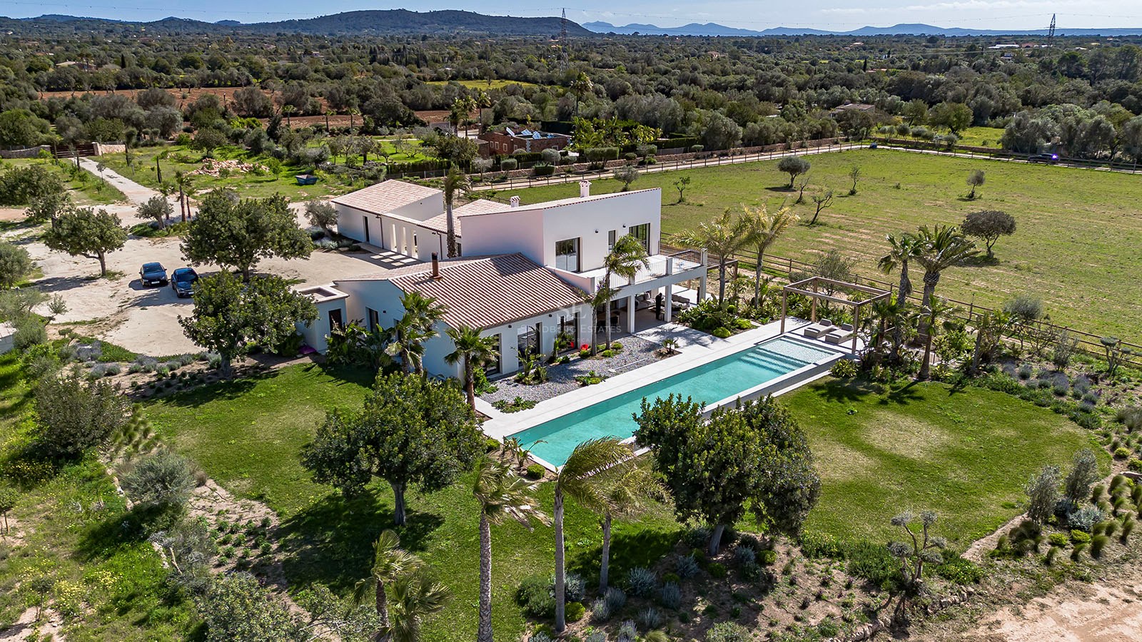 Aerial view of a large white villa with a long swimming pool, surrounded by green lawns and palm trees under a blue sky.
