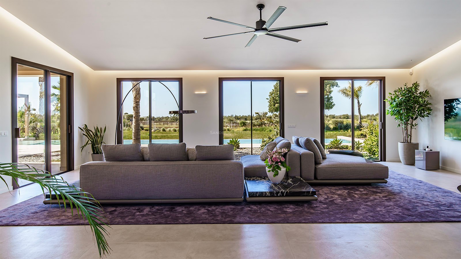 Modern living room with grey sofas, purple rug, and floor-to-ceiling glass doors opening to a lush garden.