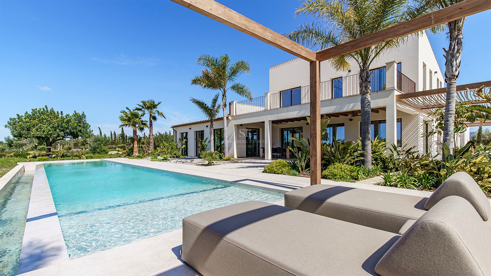 Luxury villa exterior featuring a turquoise pool, sun loungers under a wooden pergola, and palm trees.