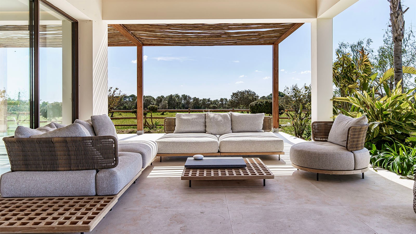 Modern lounge furniture on a shaded stone terrace under a wooden pergola overlooking a Mediterranean landscape.
