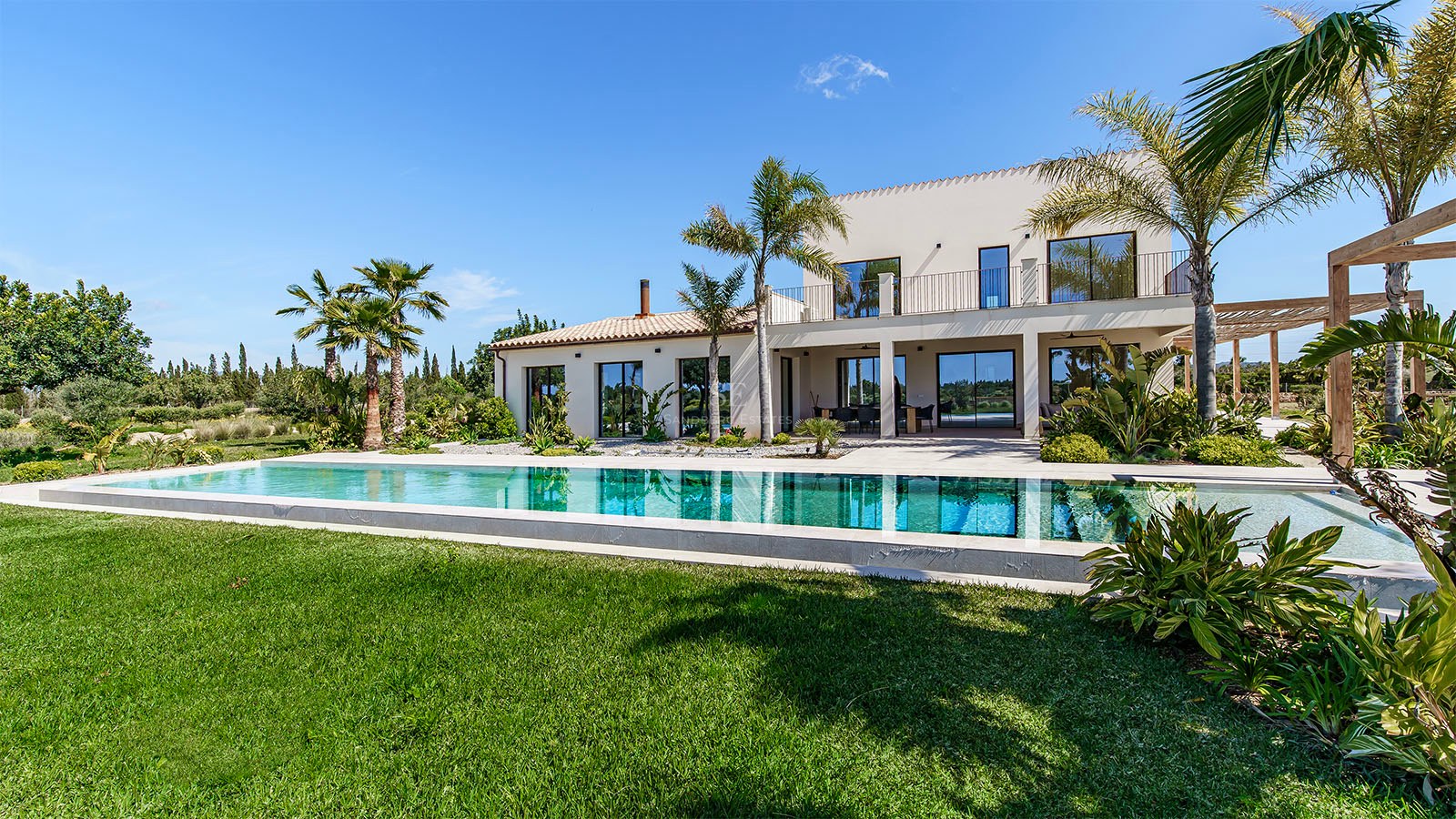Contemporary white villa featuring a long rectangular swimming pool, manicured green lawn, and palm trees under a clear blue sky.