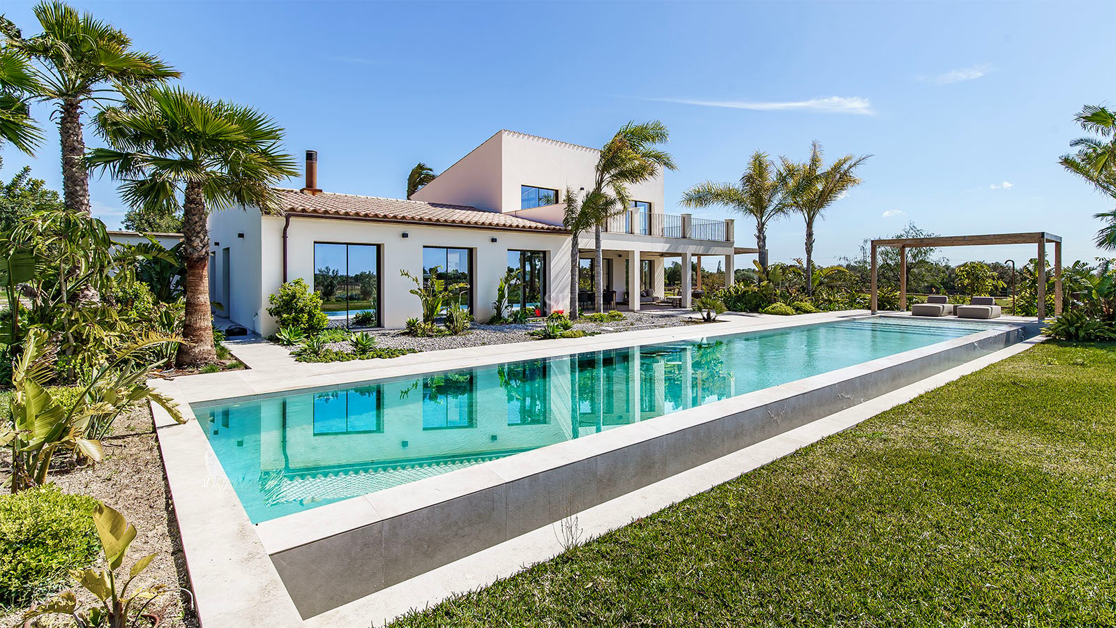 Contemporary white villa with a large rectangular swimming pool, manicured lawn, and palm trees under a clear blue sky.