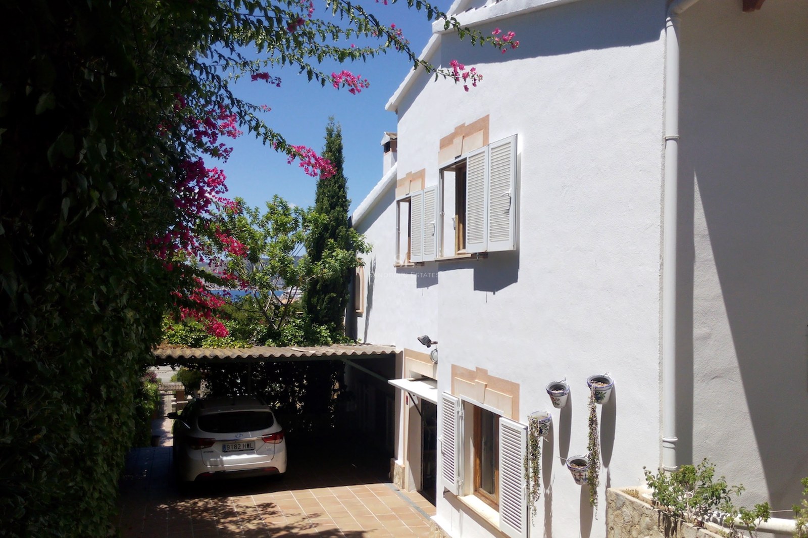 Villa à deux étages aux murs blancs, volets blancs et bougainvilliers, avec un parking couvert en terre cuite.