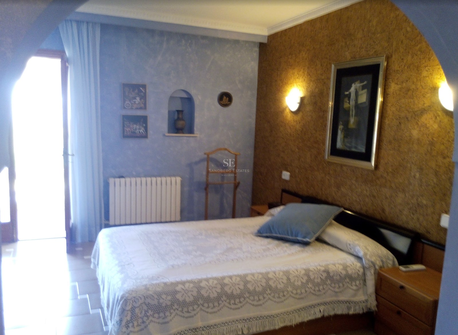 Chambre avec couvre-lit en dentelle blanche, mur texturé bleu et lumière naturelle provenant d'une porte.