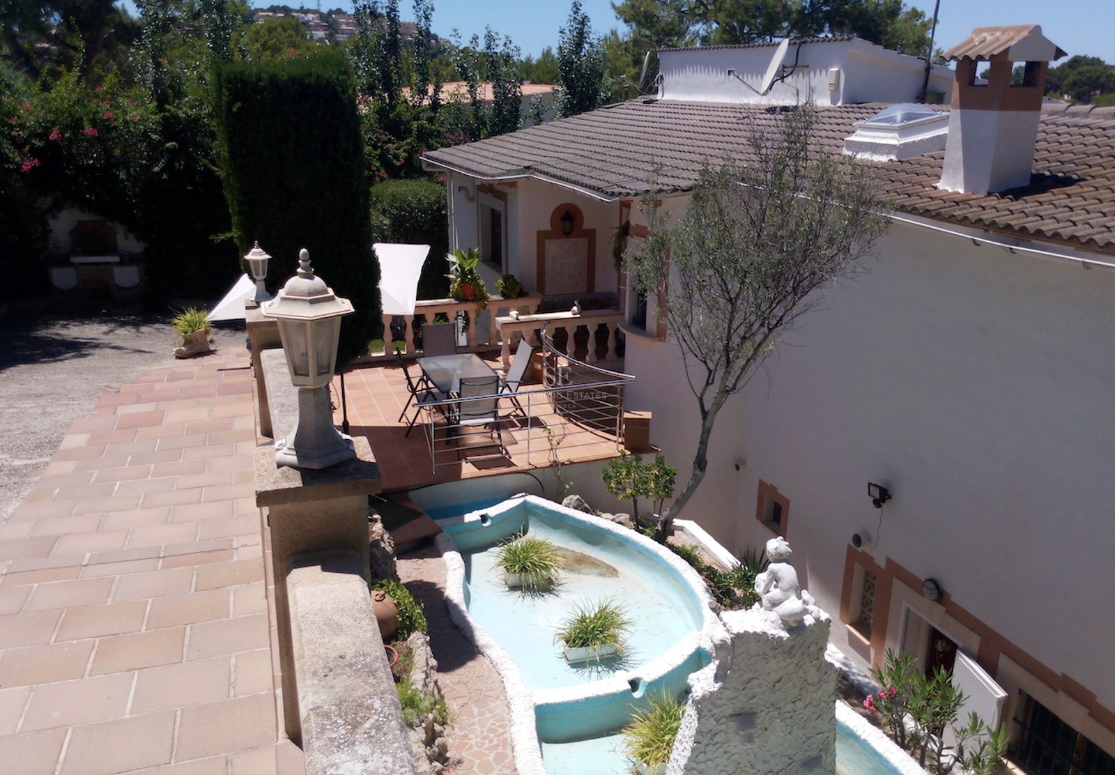Terrasse en terre cuite avec table à manger en verre, façade blanche et fontaine décorative sous la lumière du jour.