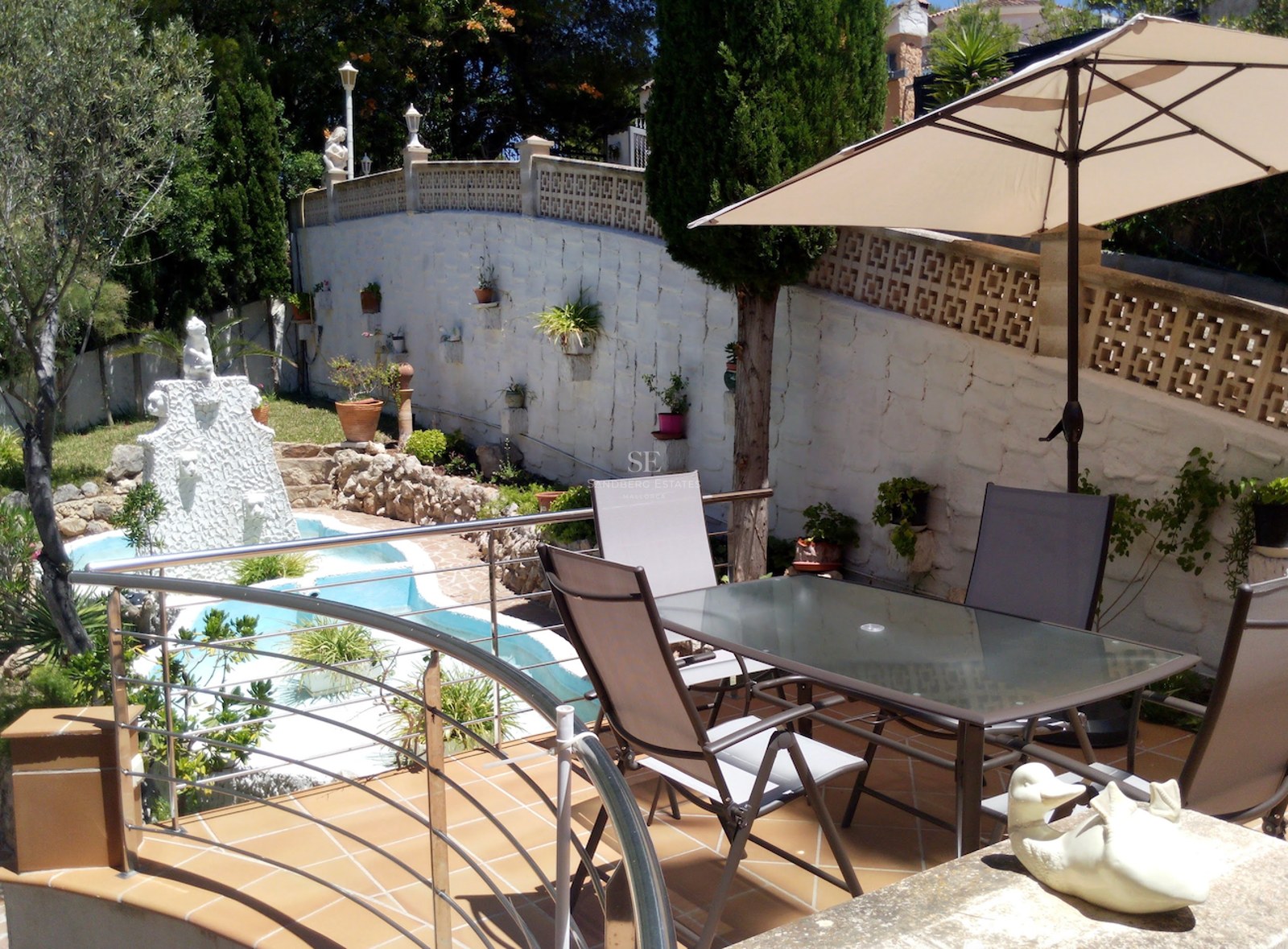 Terrasse avec table en verre et parasol surplombant une fontaine de jardin et des arbres méditerranéens.