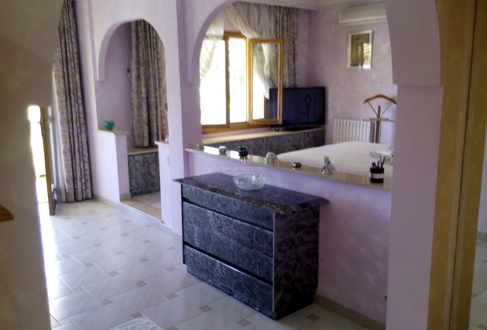 Chambre de maître aux tons lilas avec des arches architecturales, commode en marbre et sol en carrelage clair.
