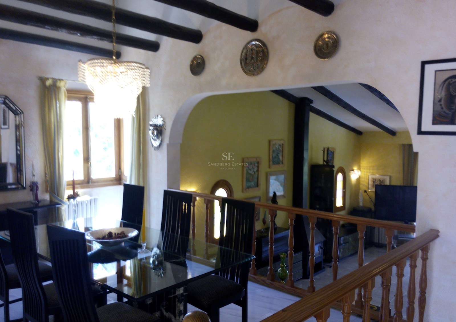 Salle à manger avec table en verre et poutres sombres, avec une balustrade en bois surplombant un salon en contrebas.