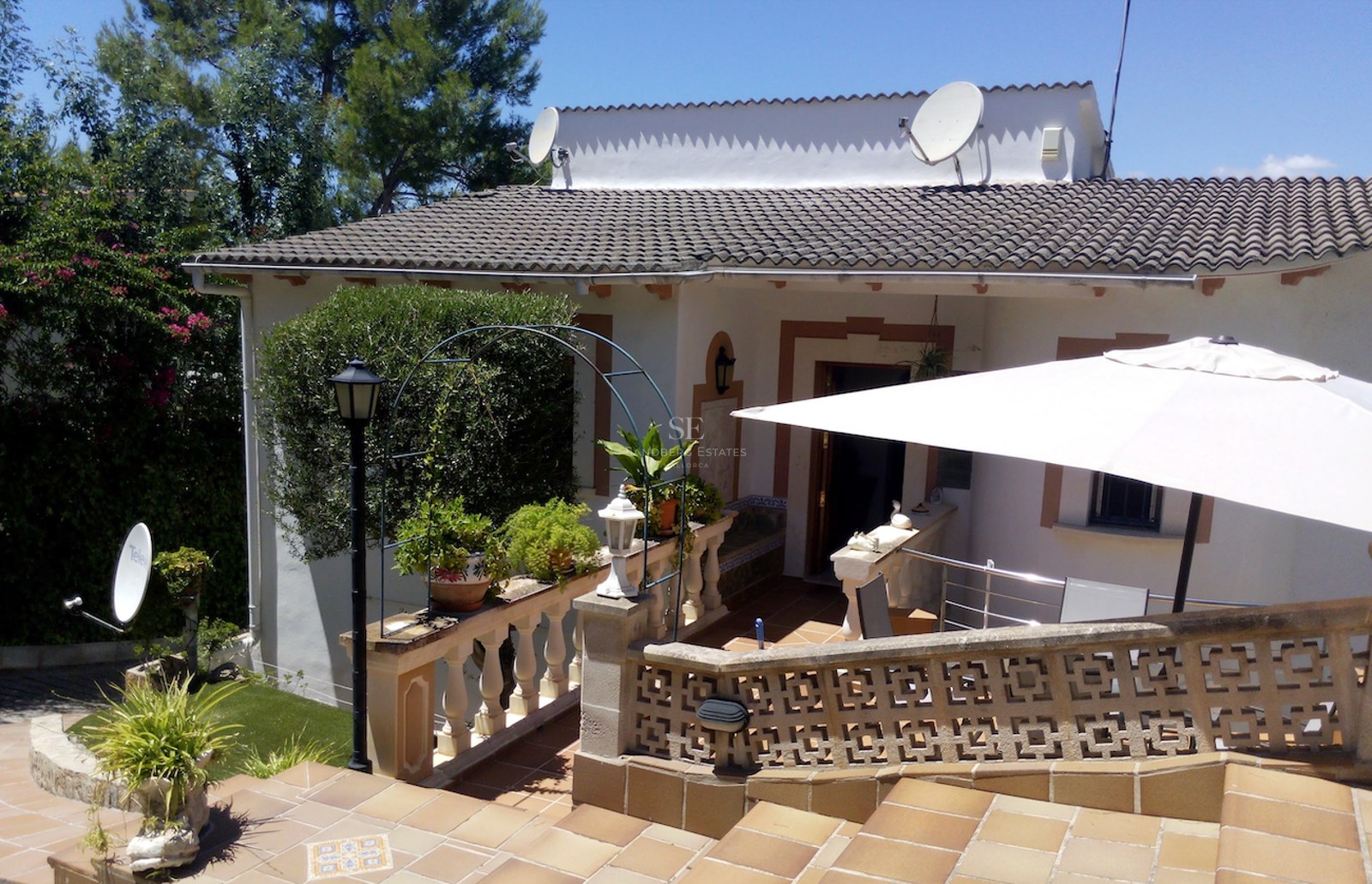 Terrasse en terre cuite avec balustrade en pierre, parasol blanc et plantes vertes sous un ciel bleu.