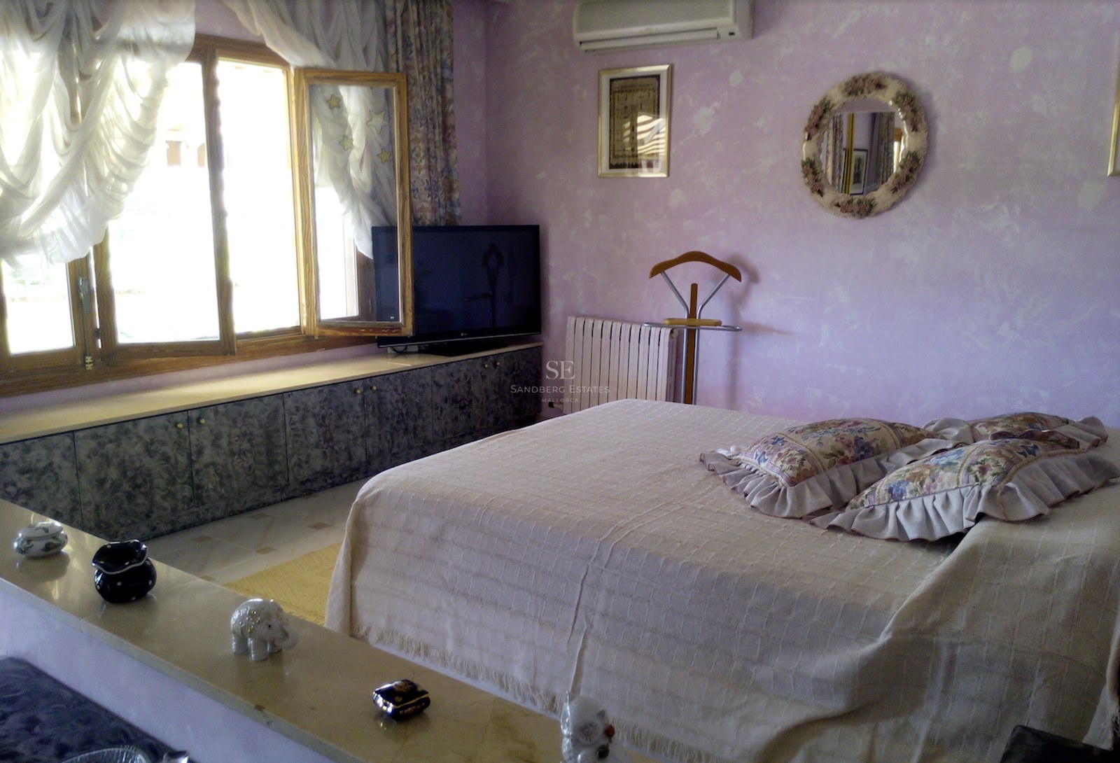 Une chambre aux murs lilas avec un grand lit, des coussins à fleurs et de grandes fenêtres en bois à la lumière du jour.