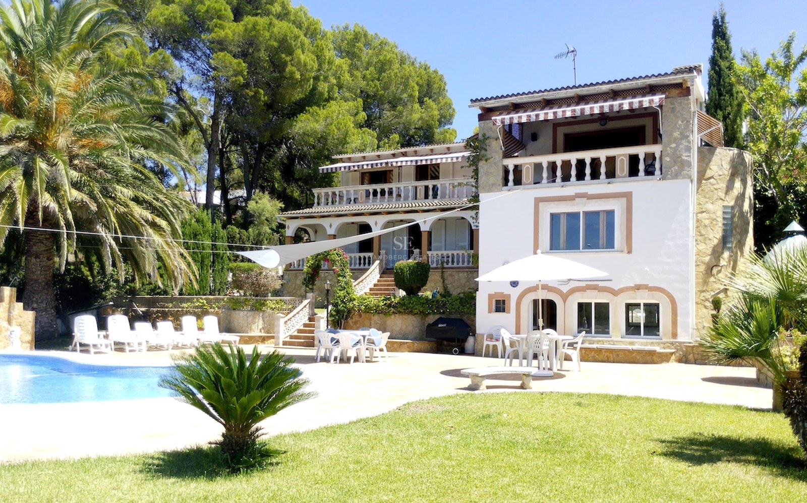 Vue extérieure d'une villa blanche à plusieurs niveaux avec accents en pierre, piscine, palmiers et terrasses.