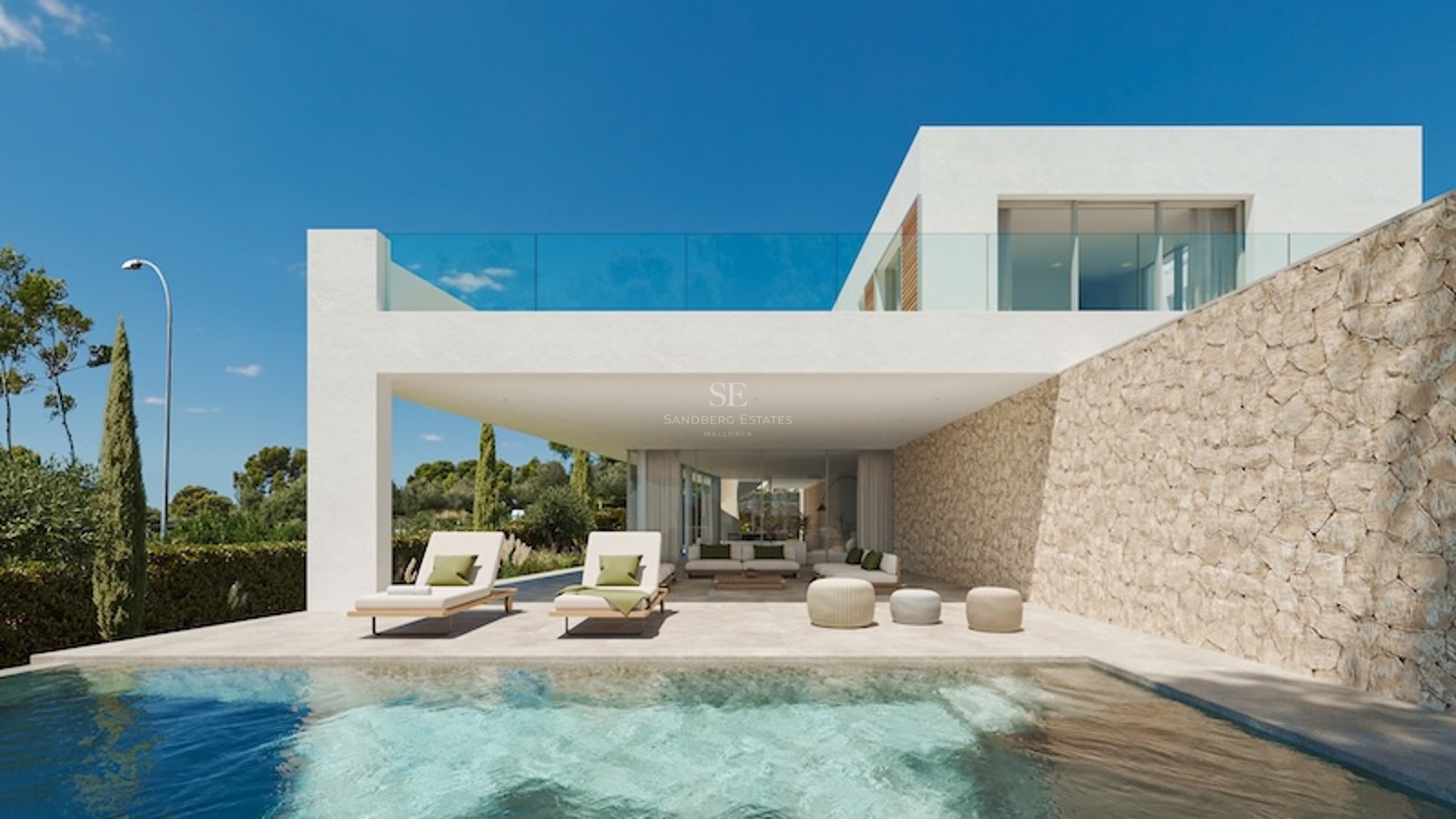 Vy över en pool mot en vit modern villa med skuggad terrass, stenvägg och solstolar under en blå himmel.
