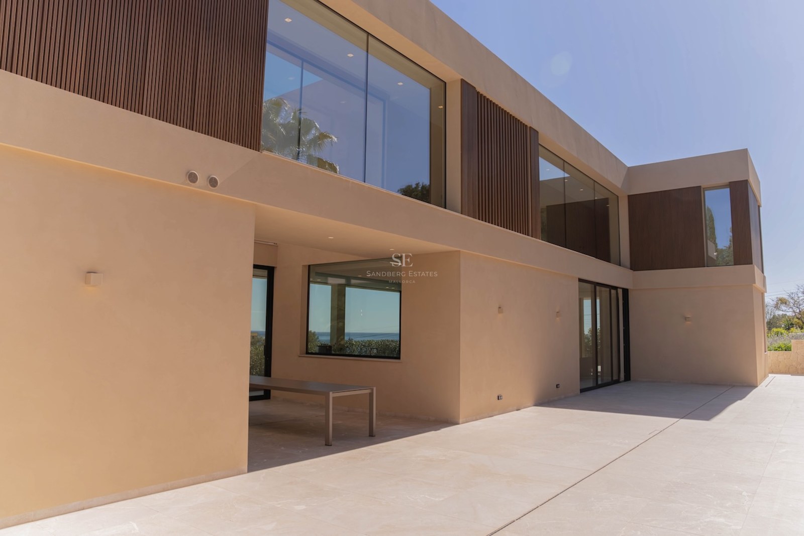 Façade d'une villa moderne beige avec de grandes vitres, des détails en bois et une terrasse en pierre.