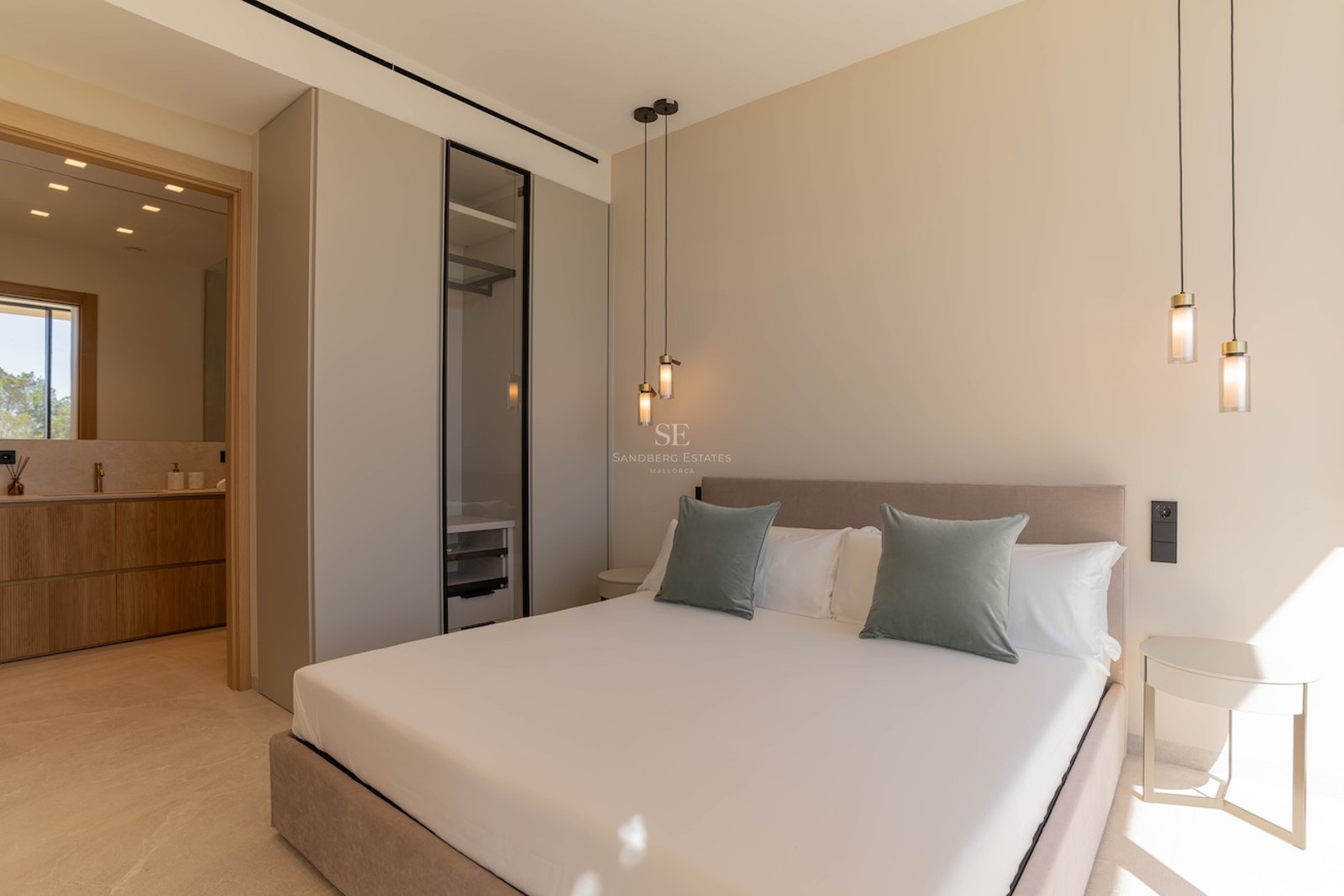 Chambre principale minimaliste avec grand lit, coussins vert sauge, suspensions design et vue sur la salle de bain.