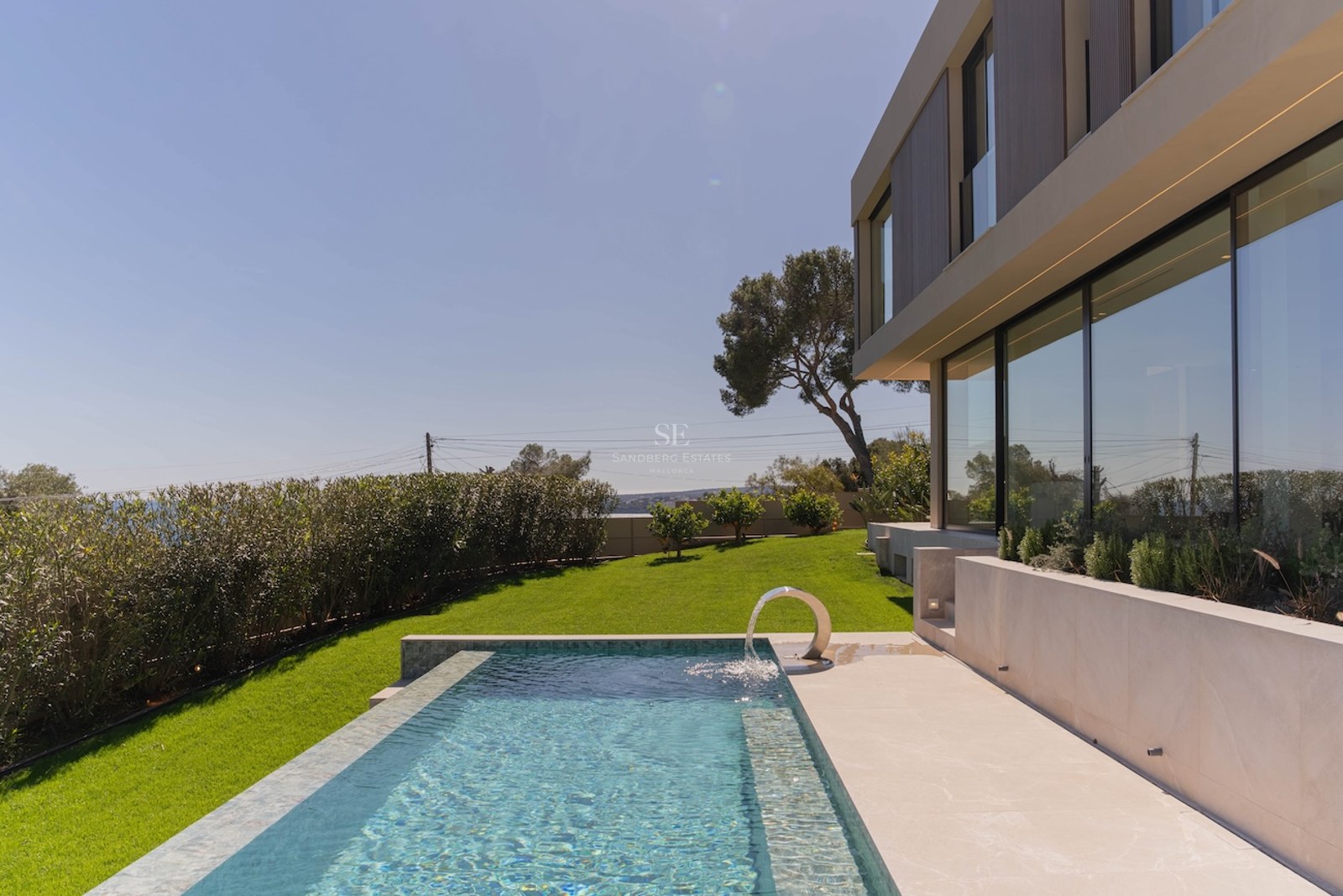 Piscine rectangulaire avec fontaine à côté d'une villa moderne aux grandes baies vitrées et pelouse face à la mer.