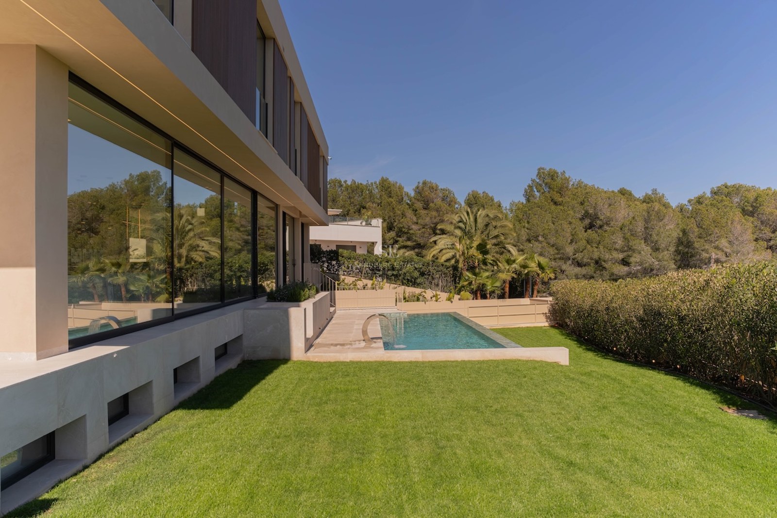 Vue latérale d'une villa moderne avec baies vitrées, piscine rectangulaire et pelouse devant une forêt dense.
