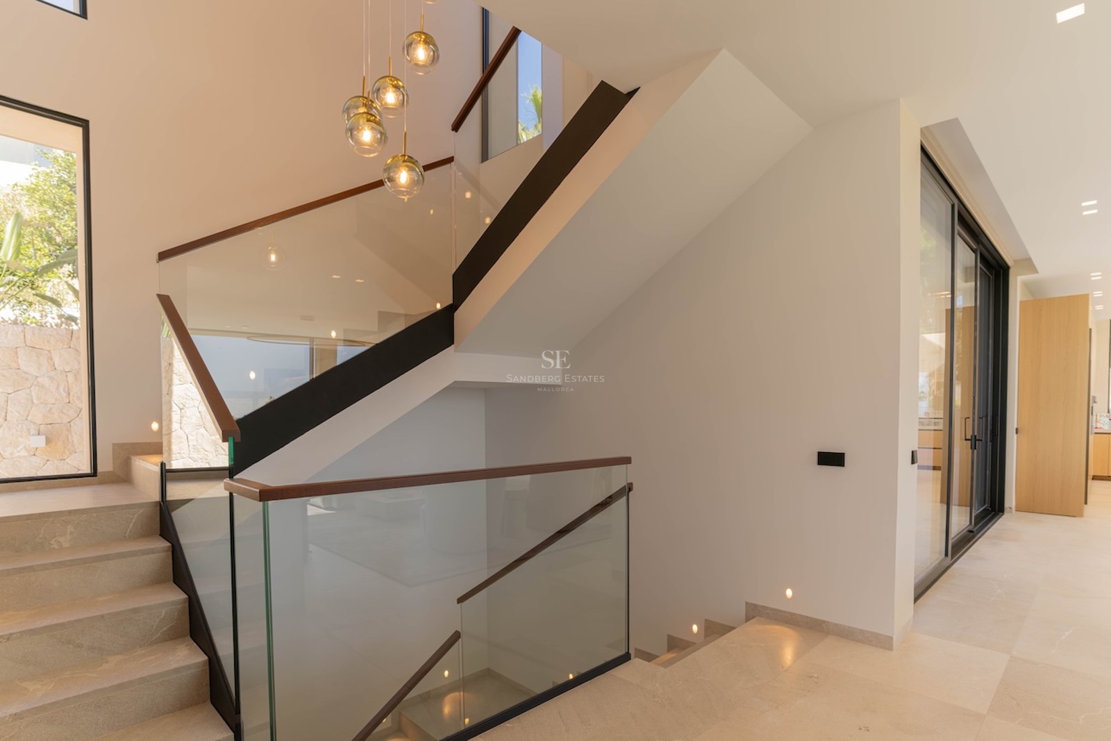 Escalier intérieur minimaliste en pierre avec balustrades en verre et suspensions dorées dans une villa lumineuse.