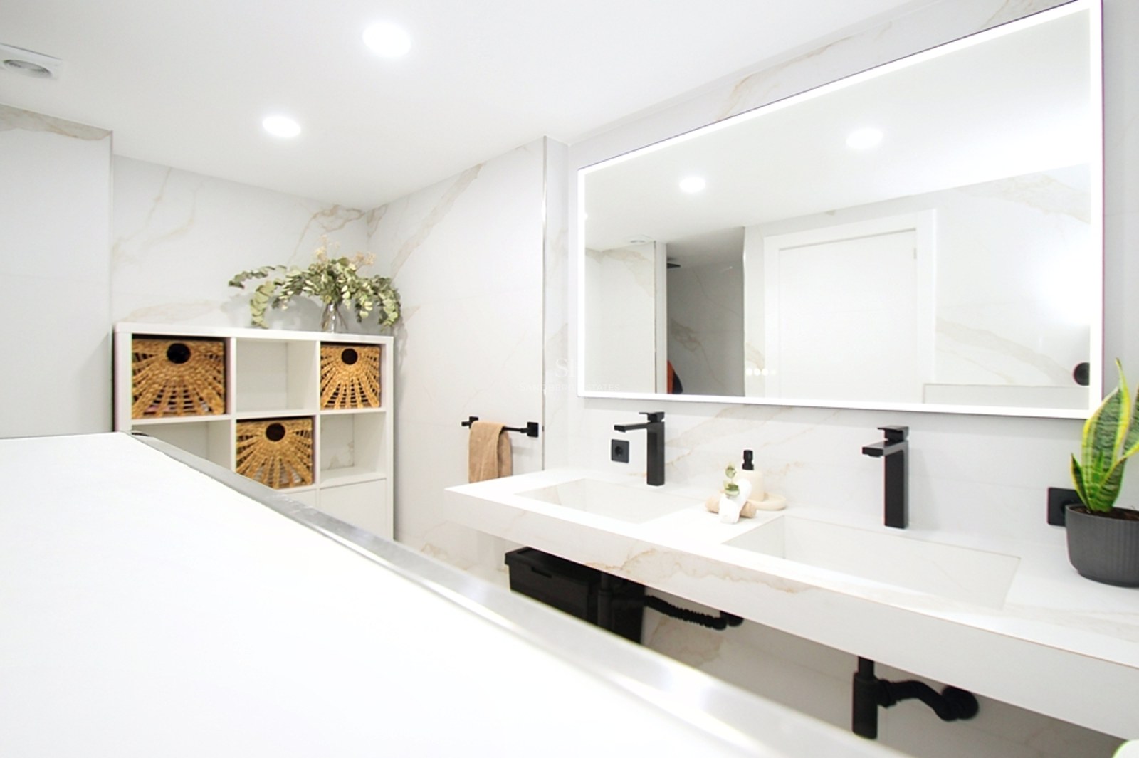 Baño moderno luminoso con lavabo doble de mármol, grifería negra mate y un gran espejo retroiluminado.