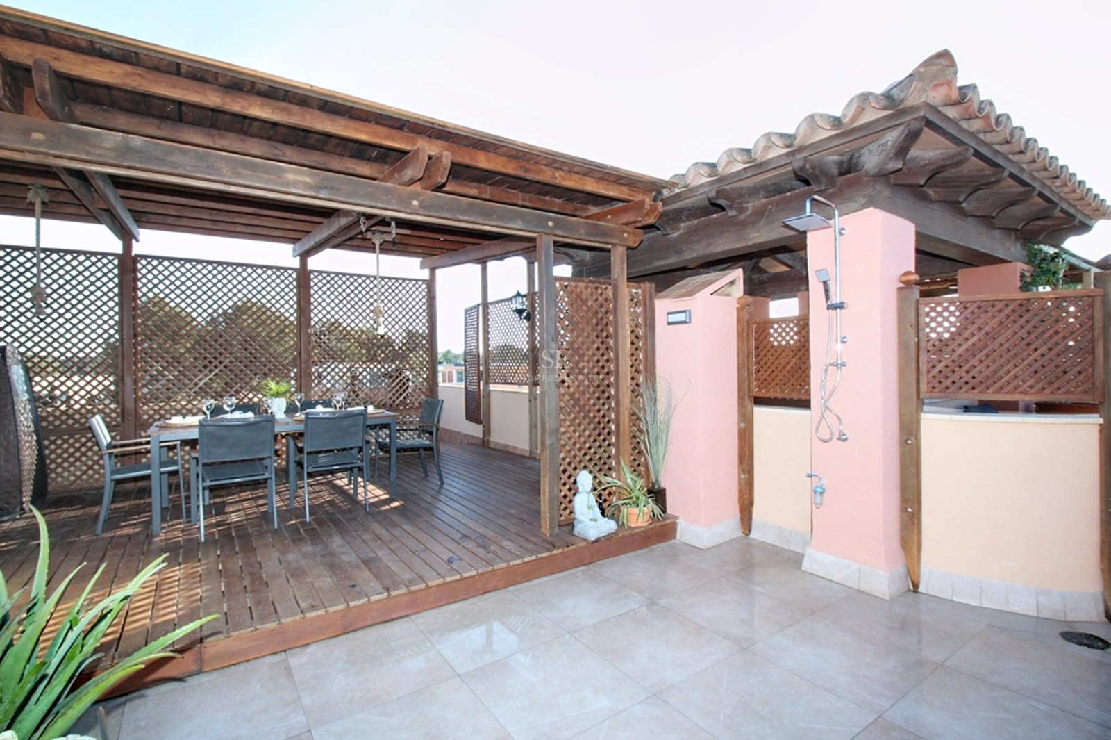 Terraza con pérgola de madera, mesa de comedor para seis personas y ducha exterior integrada junto a muros rosados.