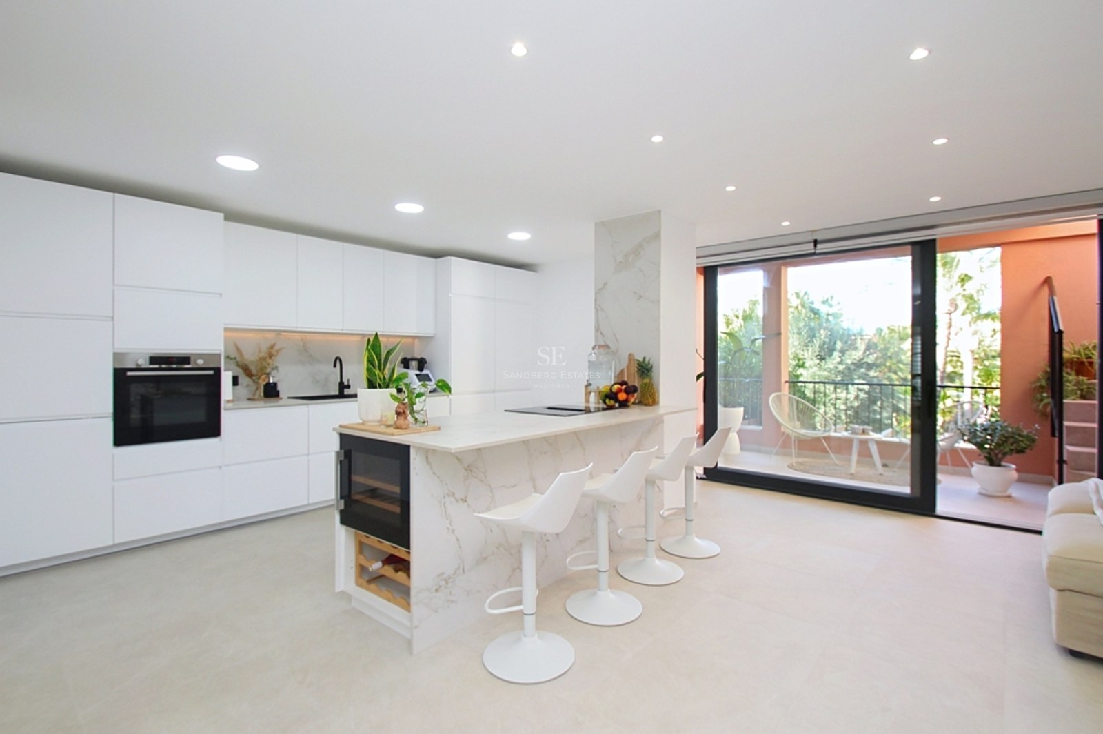 Cocina blanca minimalista con isla de mármol, vinoteca y puertas de cristal que dan a una terraza soleada.