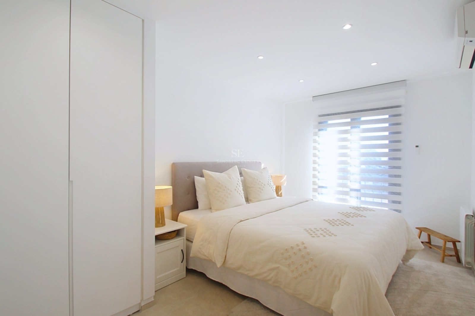 Dormitorio moderno de tonos neutros con cama doble, armarios blancos empotrados y luz natural a través de estores.