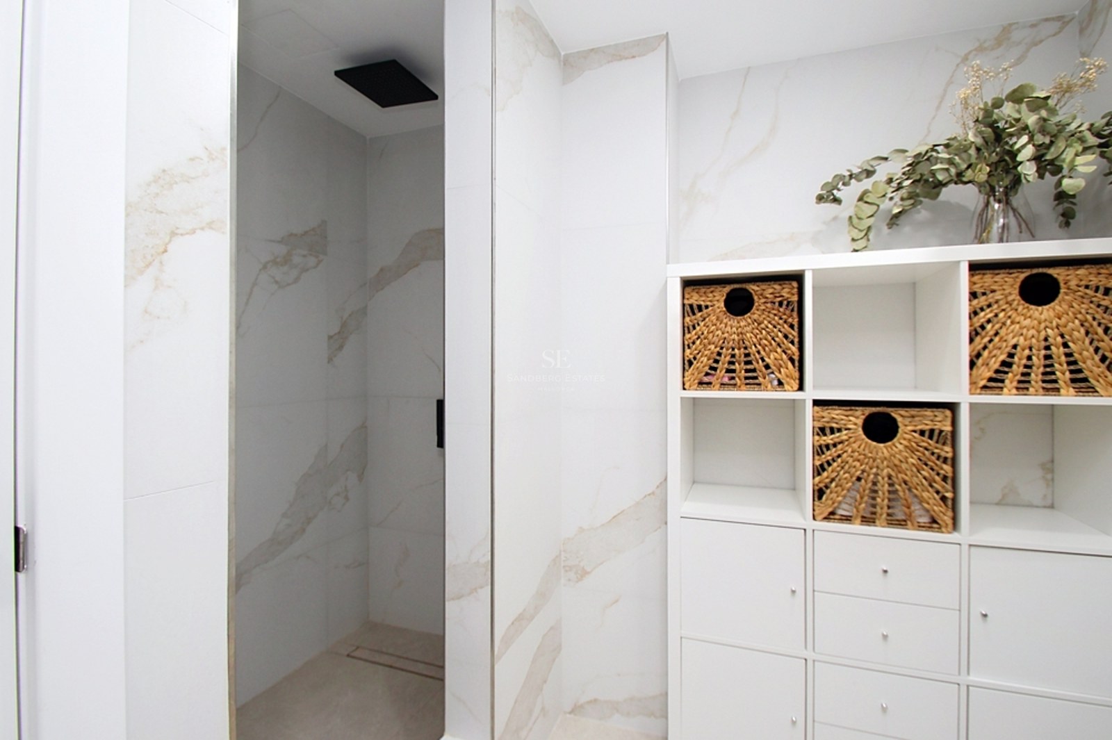 Baño contemporáneo con ducha de efecto mármol, cabezal de lluvia negro y estantería blanca con cestas de mimbre.