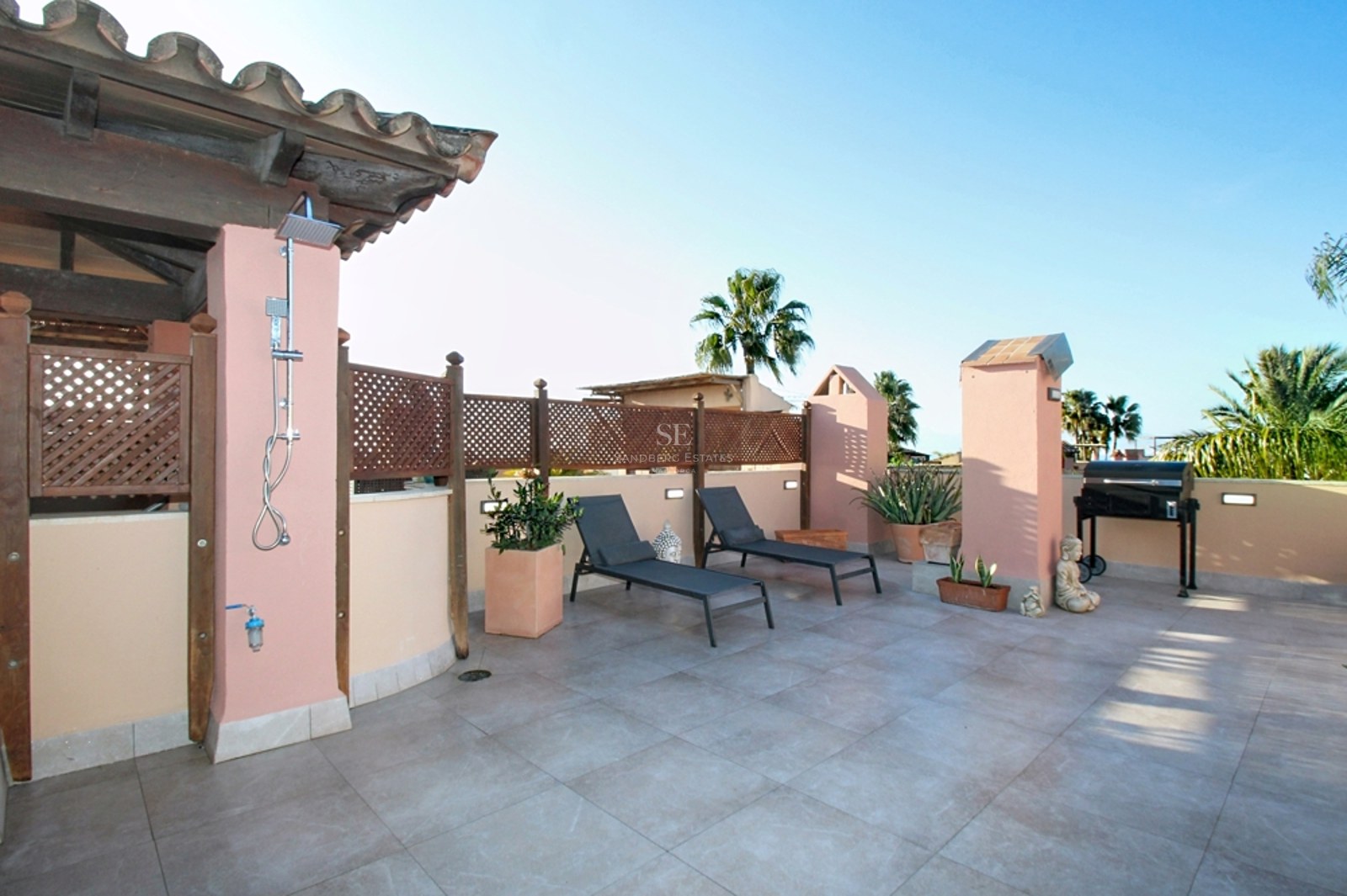 Amplia terraza mediterránea con tumbonas, ducha exterior, barbacoa y suelos de piedra clara bajo cielo despejado.