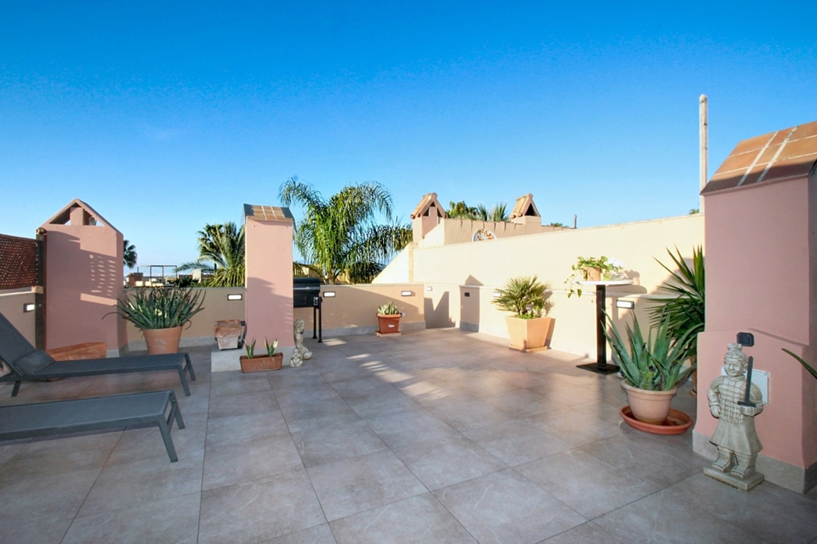 Gran terraza en azotea con azulejos beige, paredes de estuco rosa, tumbonas, plantas y cielo azul despejado.