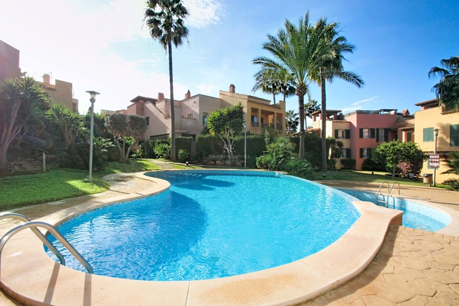 Piscina de forma irregular rodeada de jardines tropicales y edificios mediterráneos bajo un cielo despejado.