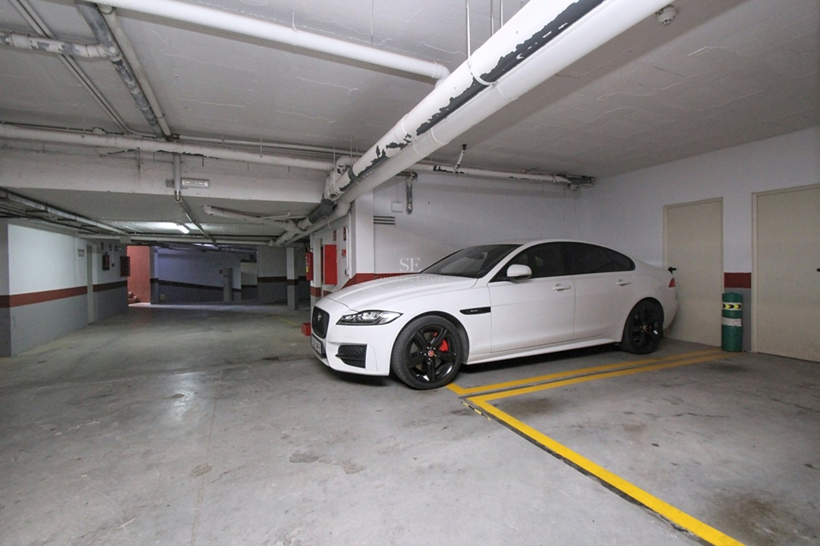 Coche blanco aparcado en un garaje subterráneo iluminado con suelo de hormigón y señalización amarilla.